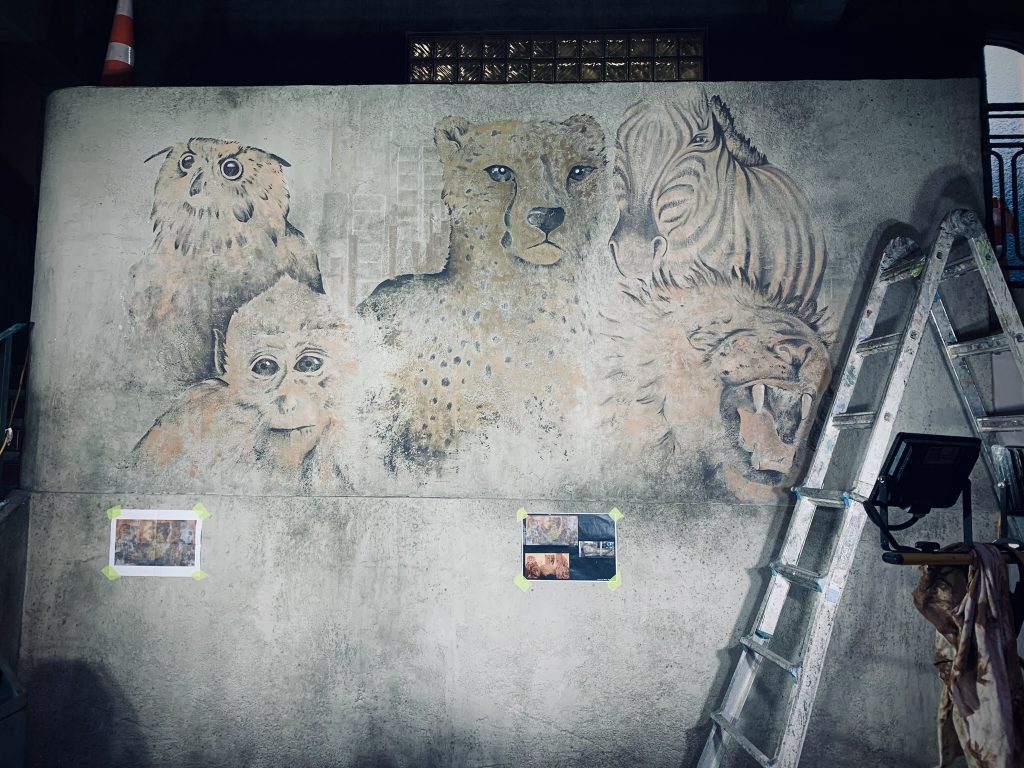 Wall mural featuring realistic animal portraits including an owl, monkey, cheetah, and zebra, painted in earthy tones on a concrete wall.